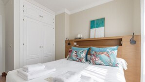 Ein ordentlich bezogenes Bett mit weißen Leinentextilien und zwei Zierkissen, ein Holz-Kopfteil, ein weißer Kleiderschrank und ein an der Wand hängendes Kunstwerk.