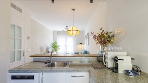 Eine moderne Küche mit Granit-Arbeitsplatte, Edelstahl-Spüle und weißen Schränken. Im Arbeitsbereich befindet sich ein eingebauter Geschirrspüler, ein Toaster und eine Kaffeemaschine. Über der Spüle hängt eine Leuchte, und auf der Arbeitsplatte steht eine Vase mit Blumen.