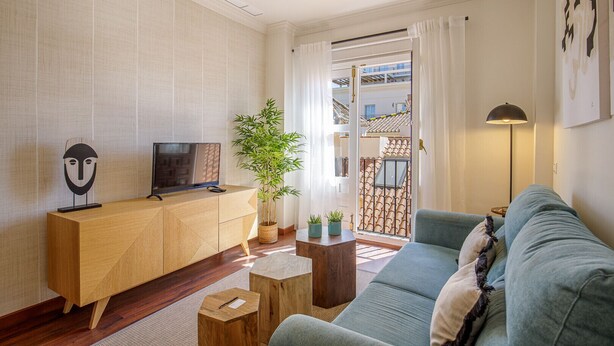 Ein Wohnzimmer mit einer Couch, einem Holz-TV-Schrank, einer Topfpflanze und Blick auf einen Balkon mit Ziegeldach.