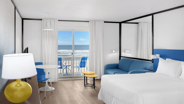 Ein Hotelzimmer mit einem Bett, einer Couch, einem kleinen Tisch, einer Lampe und Blick auf den Ozean durch die Glastür.