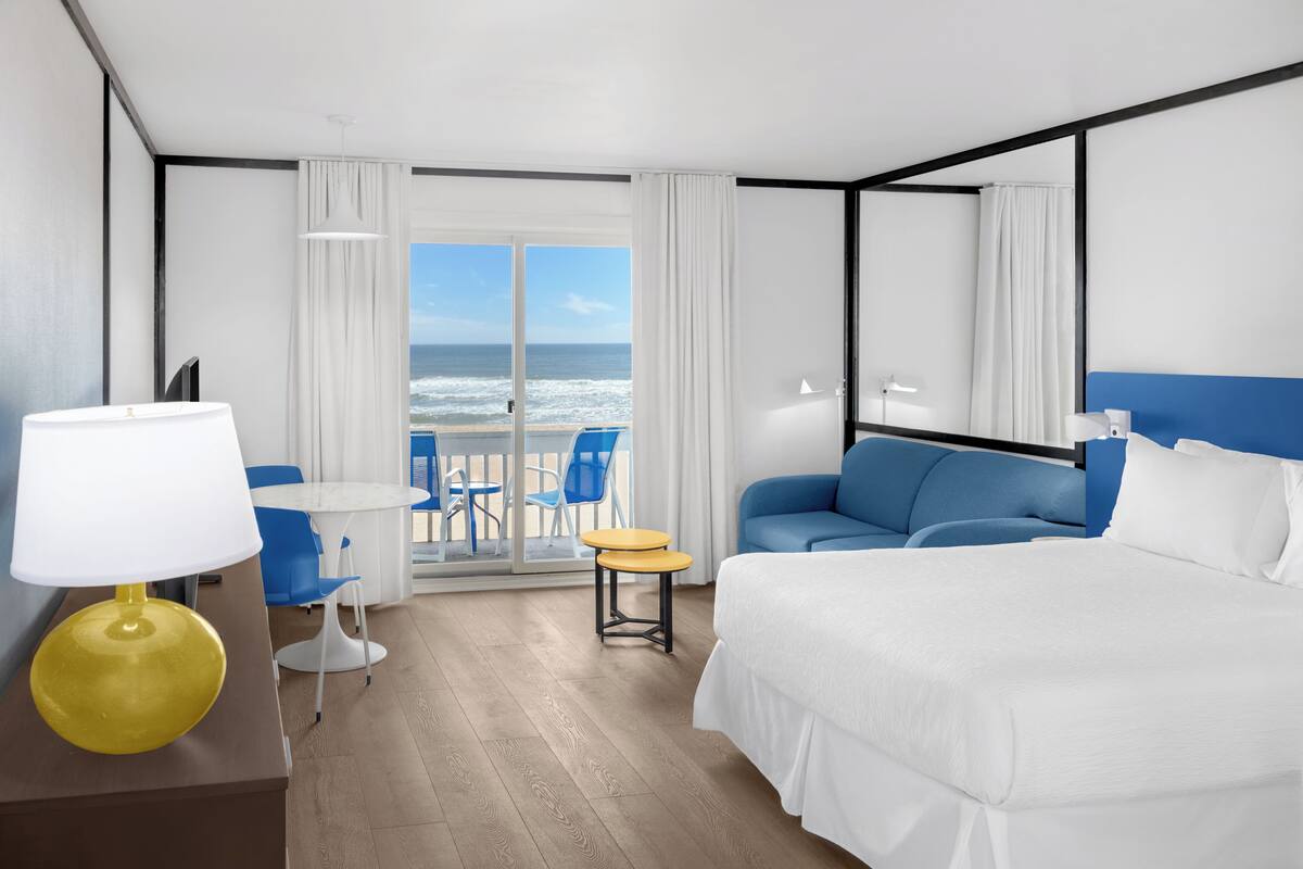 Quarto de hotel com cama, sofá, mesinha, abajur e vista para o oceano pela porta de vidro.