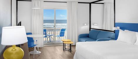 A hotel room with a bed, a sofa, a small table, a lamp, and a view of the ocean through the glass door.