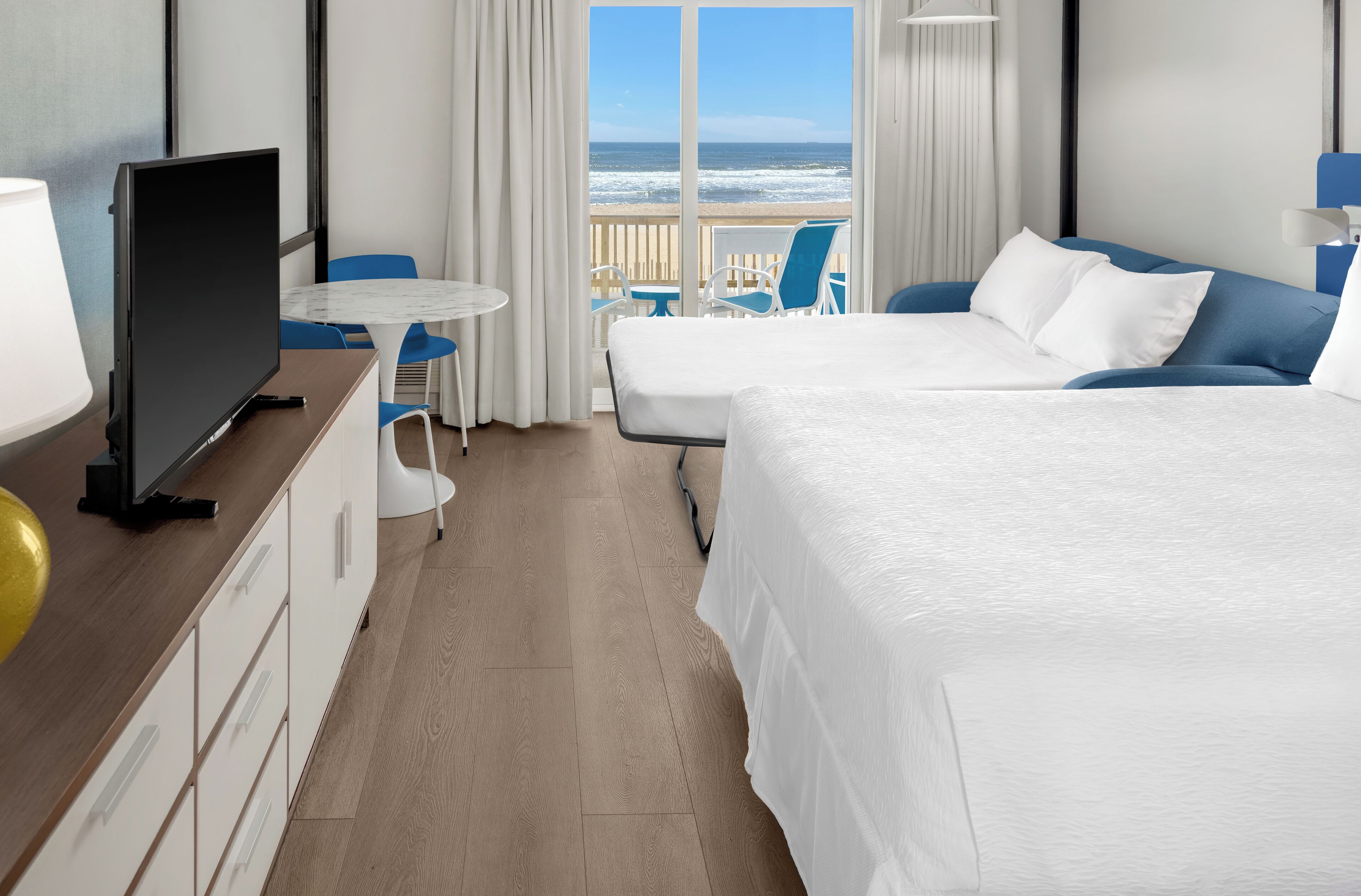 A hotel room with two beds, a TV, a desk, and a view of the beach.