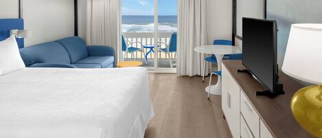 Habitación de hotel con cama, televisor, escritorio con lámpara, silla y vista al océano a través de puertas correderas.
