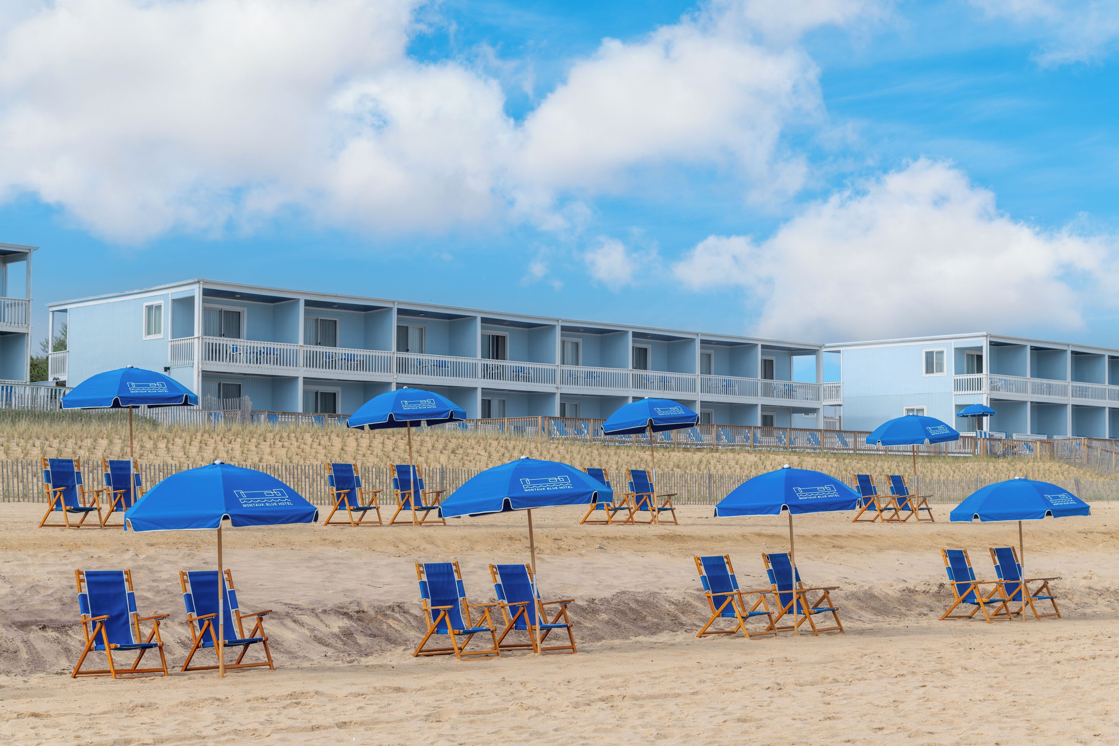A beachfront hotel with multiple blue umbrellas and beach chairs arranged in rows.