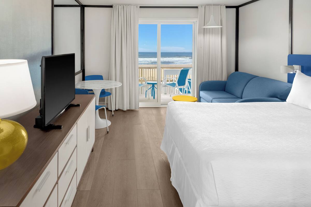 Quarto de hotel com cama, escrivaninha com televisão, mesinha e cadeiras. Tem varanda com vista para o oceano.