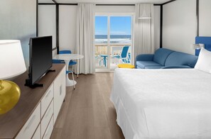 Une chambre d’hôtel avec un lit, un bureau avec une télévision, une petite table et des chaises. Il y a un balcon avec vue sur l’océan.