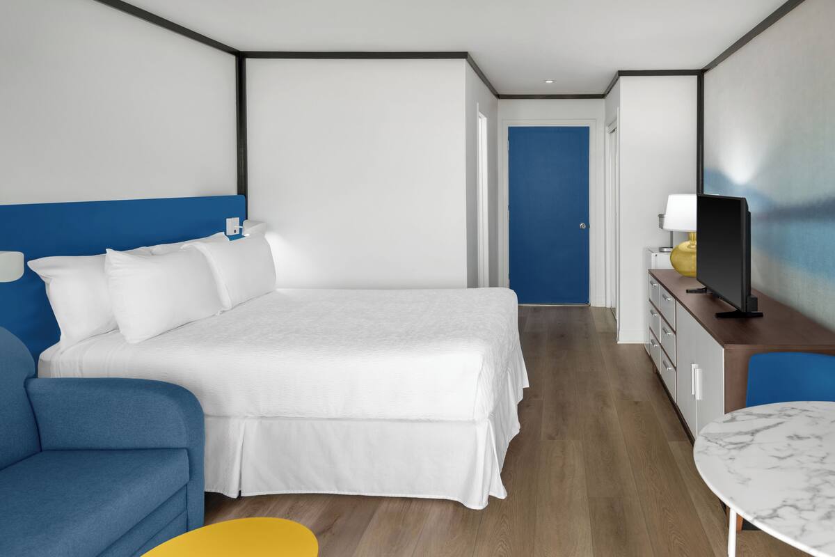 Quarto de hotel moderno com uma cama grande, um sofá azul, uma mesa pequena e uma TV em um armário de madeira.