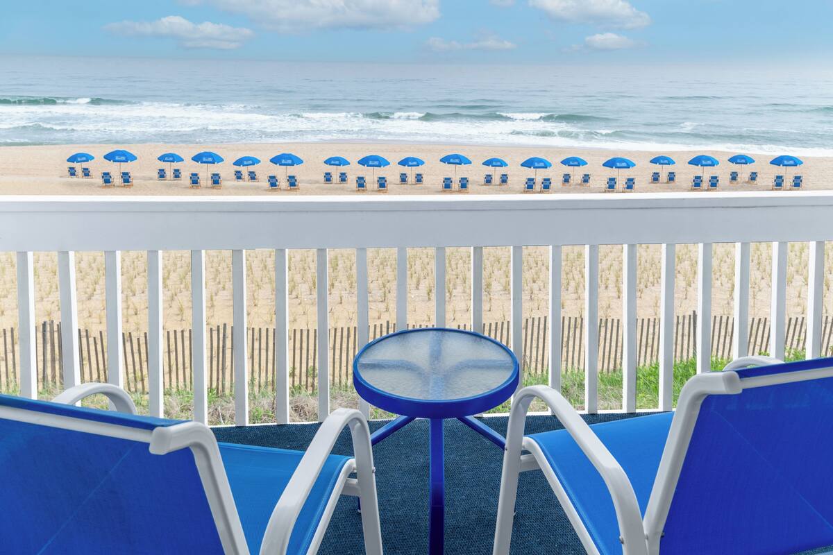 Uma varanda com vista para a praia, com cadeiras azuis, uma mesa redonda e guarda-sóis azuis.