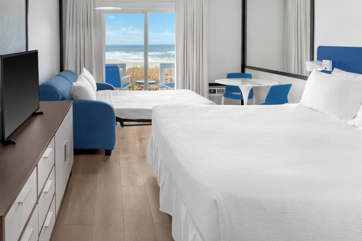 Quarto de hotel com uma cama grande, uma escrivaninha com televisão, uma pequena mesa com cadeiras e vista para o oceano.