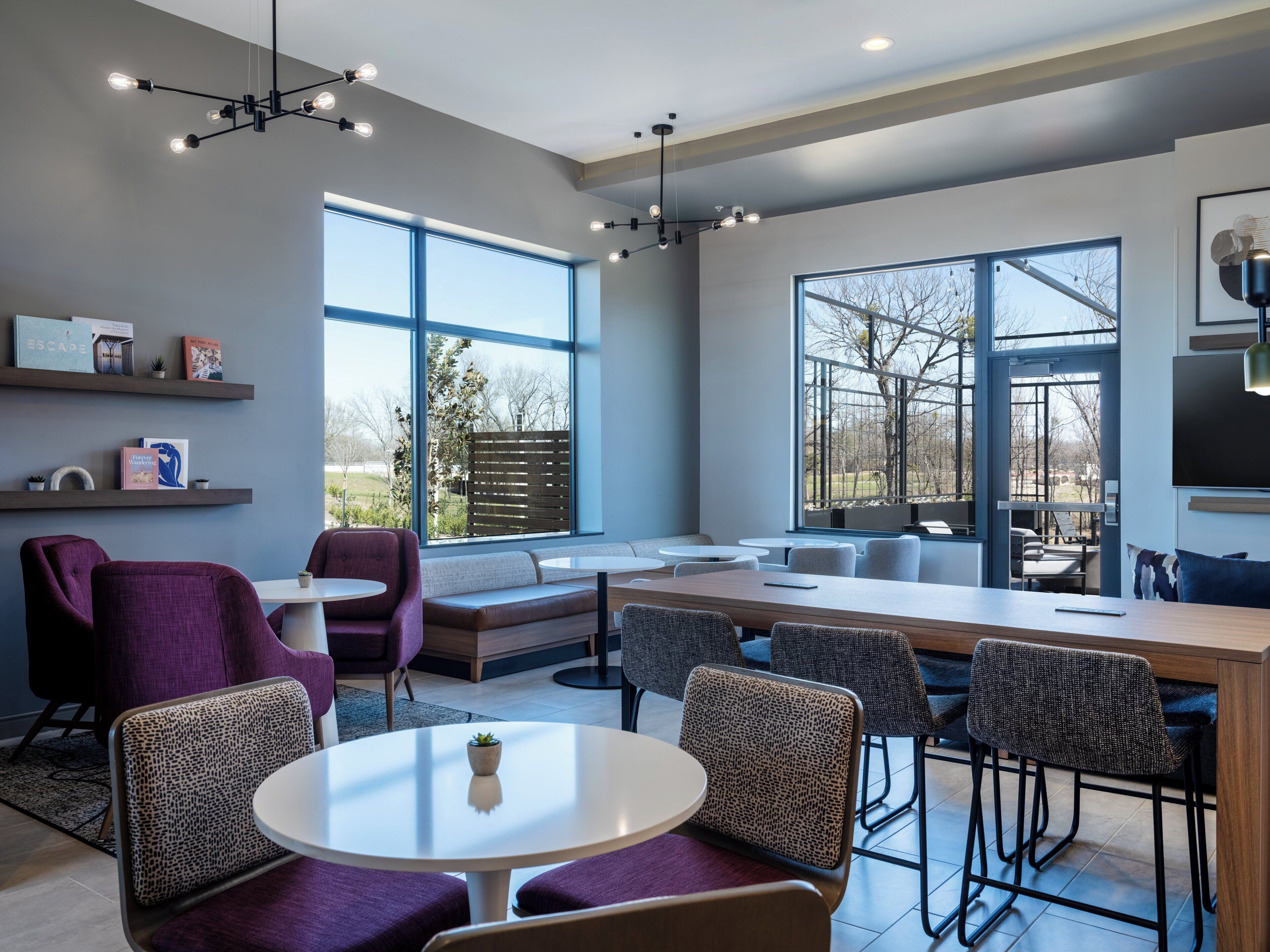 A modern dining area with a long table, multiple chairs, and a large window offering a view of the outdoors.
