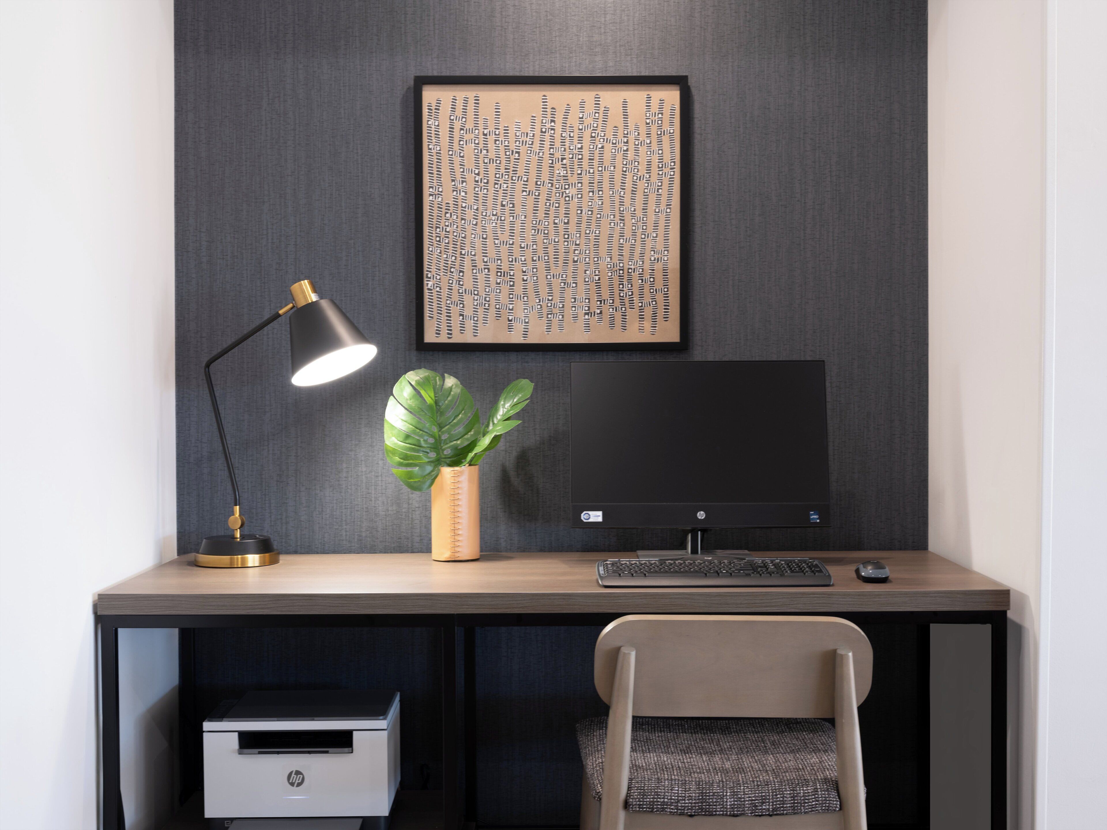 A modern office desk with a computer, keyboard, mouse, and a lamp. There is a printer and a chair in front of the desk.