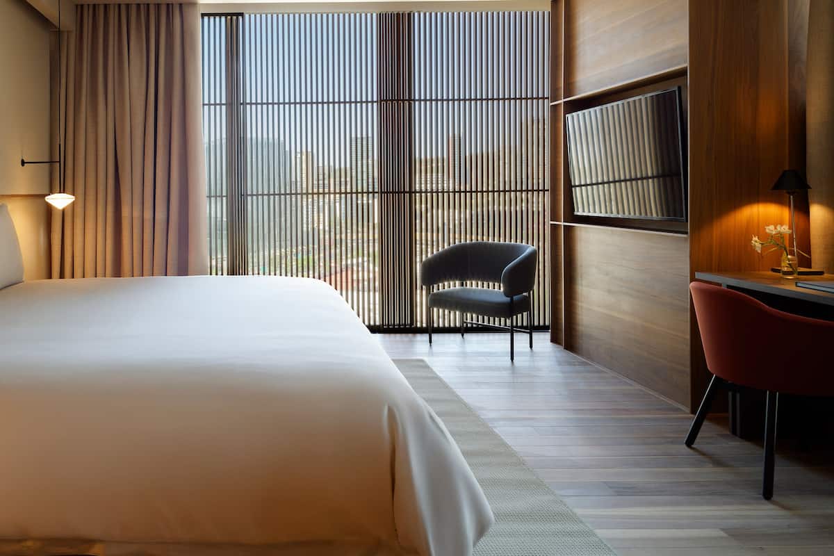 A modern hotel room with a large bed, a desk with a chair, and a view of the city through floor-to-ceiling windows.