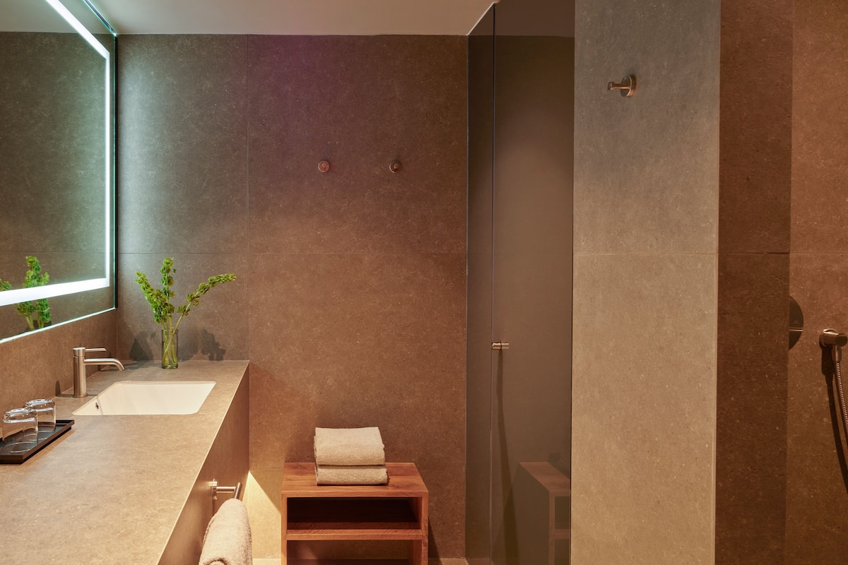 A modern bathroom with a large mirror, a sink, a shower, and a small wooden stool.