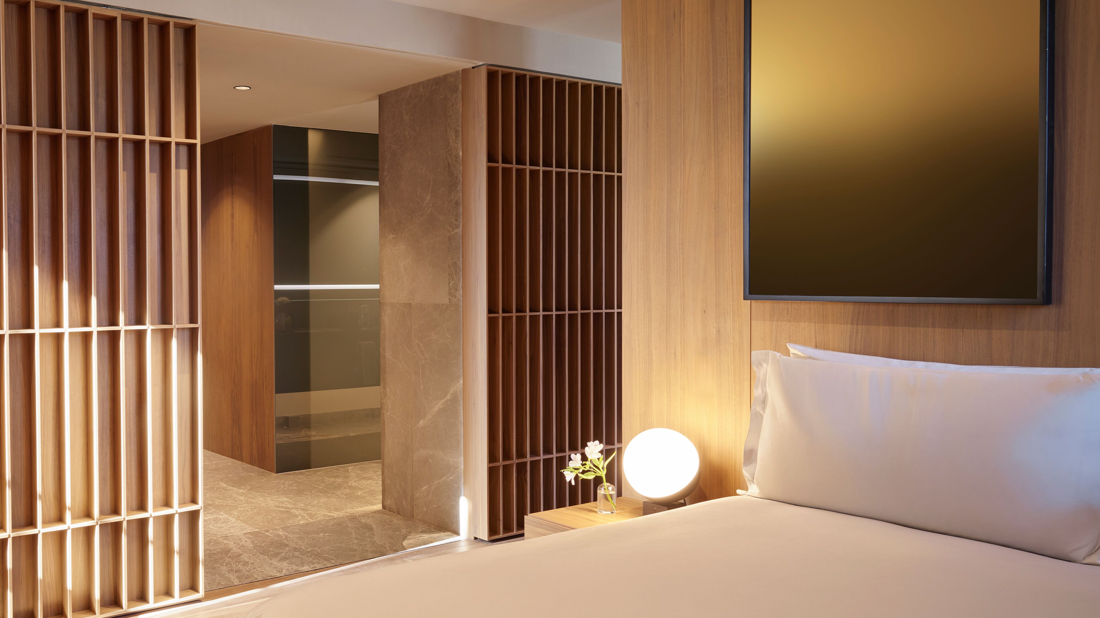 A modern hotel room with a bed, a bedside table with a lamp, a wooden partition, and a view into the hallway.