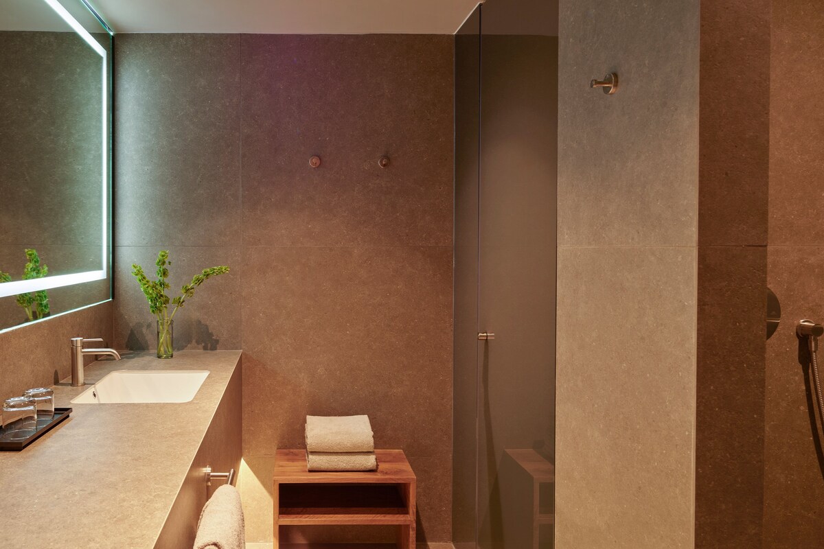 Une salle de bain moderne avec un grand miroir, un lavabo, une douche et un petit tabouret en bois.