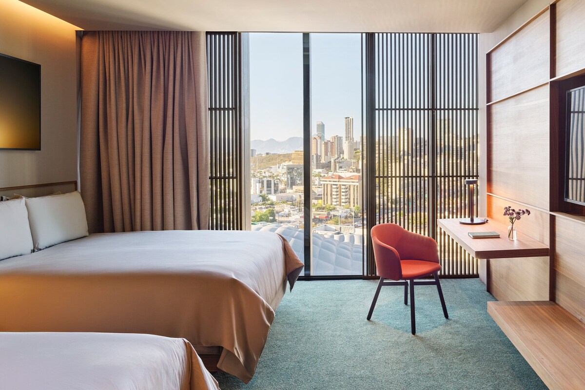 Une chambre d’hôtel avec deux lits, un bureau, une chaise et une grande fenêtre donnant sur la ville.