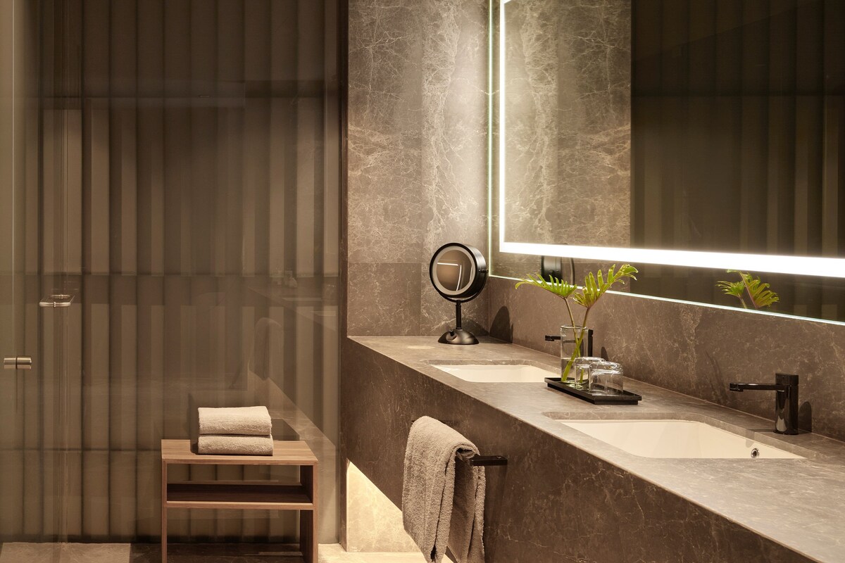 Une salle de bain moderne avec une douche à l’italienne en verre, un meuble-lavabo avec deux vasques, un grand miroir et une petite plante posée sur le plan de travail.