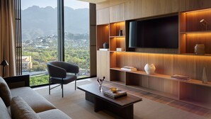 A modern living room with a large TV, wooden shelving, and a view of a cityscape through large windows. - InterContinental Presidente Monterrey by IHG (San Pedro Garza García)