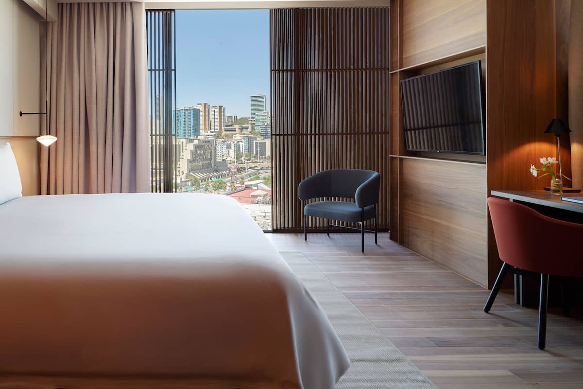 A modern hotel room with a large bed, a desk with a chair, a TV, and a view of the cityscape through large windows.