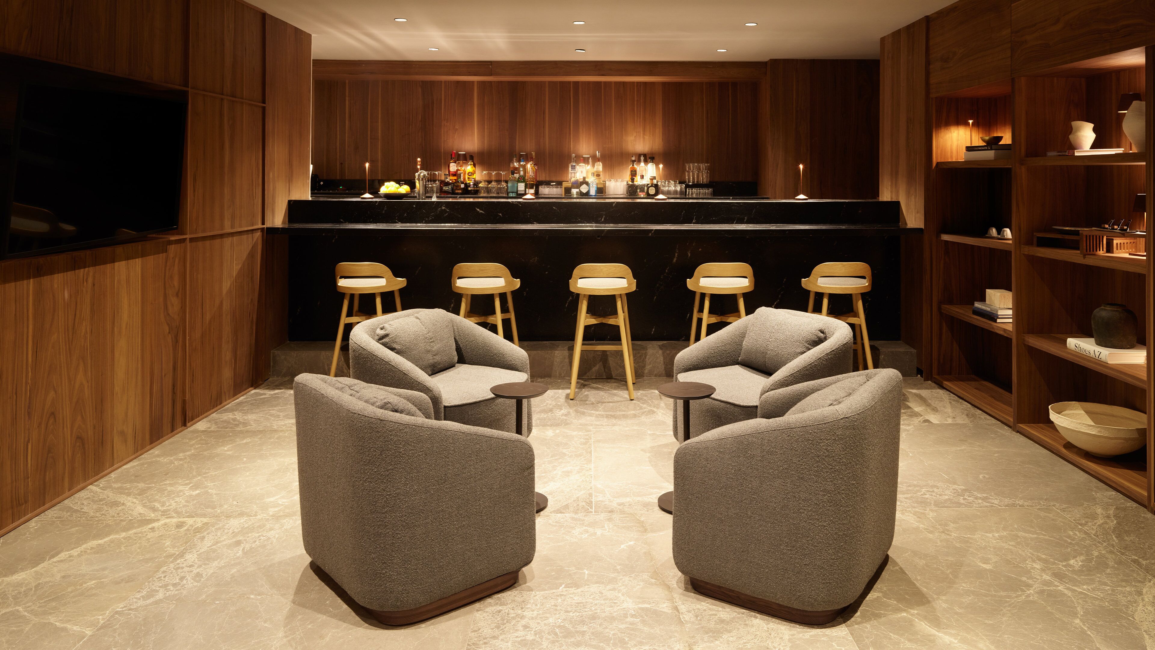 Un área de bar moderna con paneles de madera, una barra y taburetes. Hay dos sillones en primer plano.
