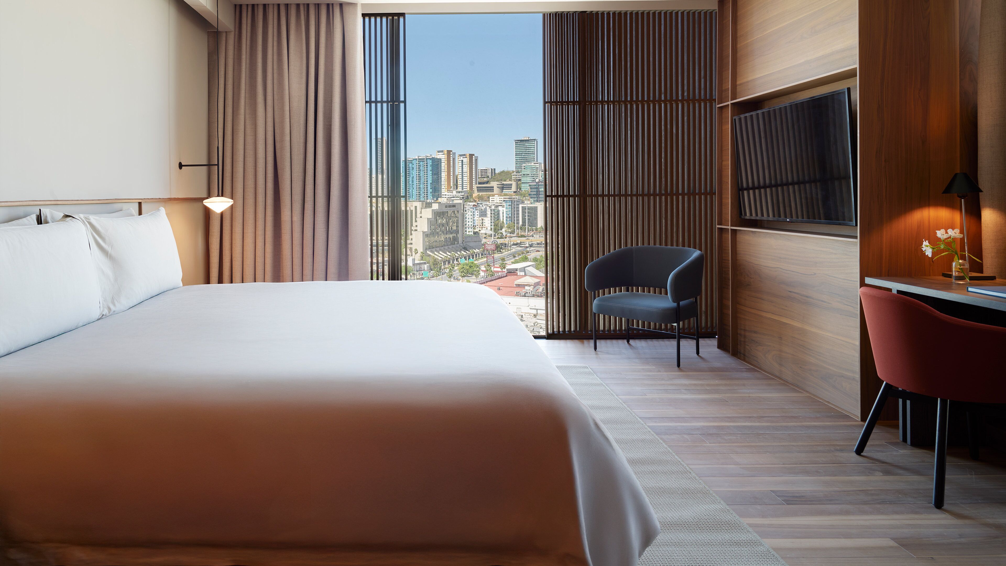 A modern hotel room with a large bed, a desk with a chair, a TV, and a view of the cityscape through large windows.
