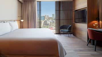 A modern hotel room with a large bed, a desk with a chair, a TV, and a view of the cityscape through large windows.