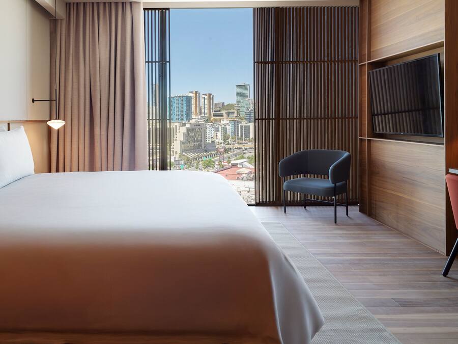 A modern hotel room with a large bed, a desk with a chair, a TV, and a view of the cityscape through large windows.
