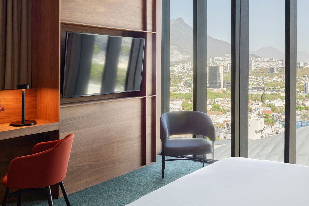 Une chambre d’hôtel dotée d’une grande fenêtre, d’un téléviseur à écran plat, d’un bureau avec une lampe, de deux chaises et d’un lit.