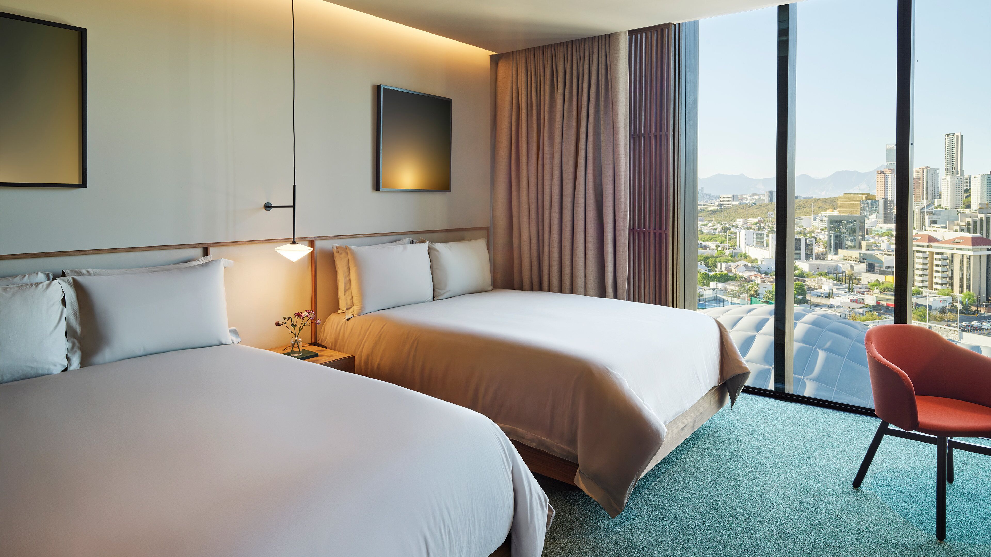 A hotel room with two beds, a desk with a lamp, a chair, and a large window with a city view.