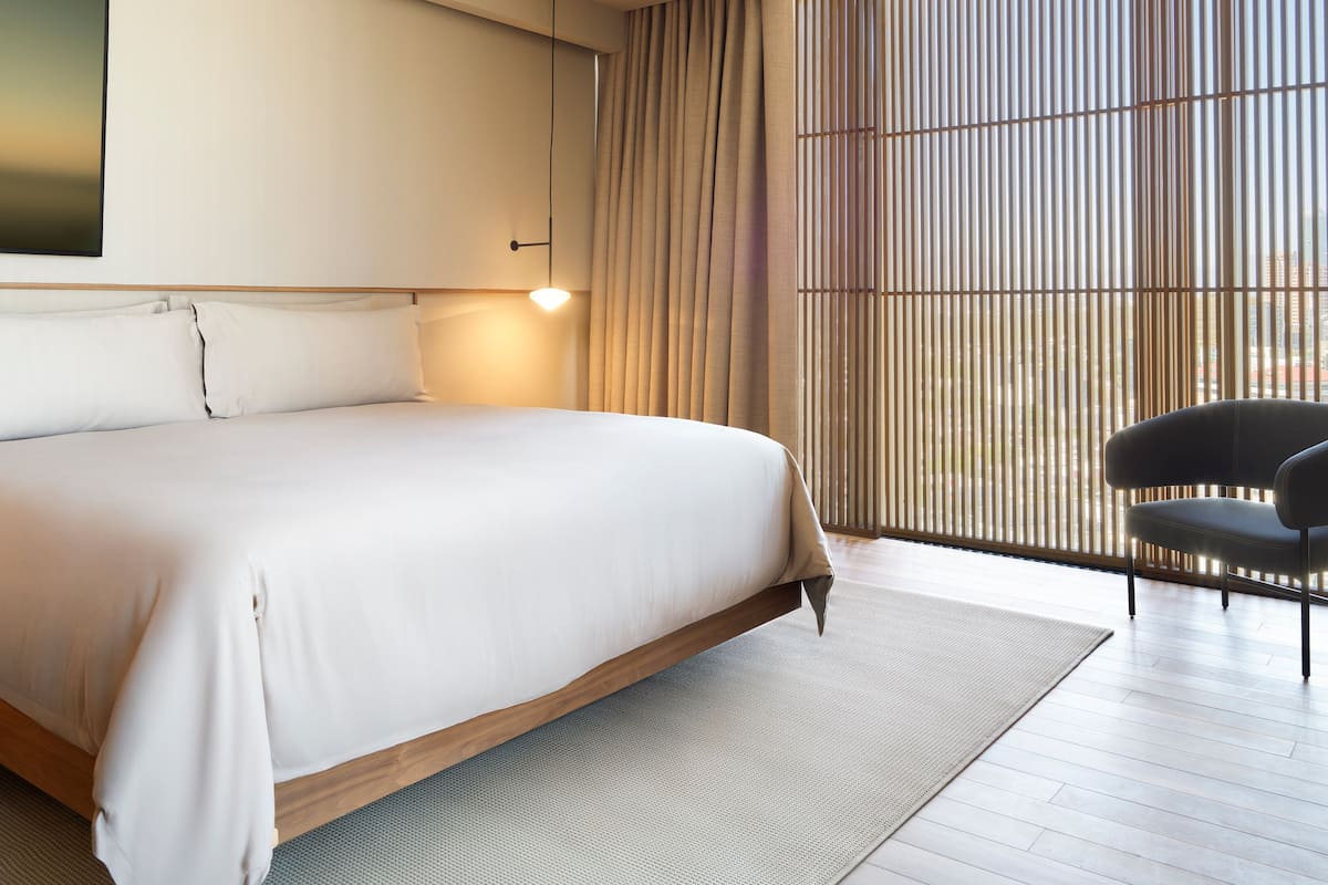 A modern hotel room with a large bed, a nightstand, a chair, and a view through floor-to-ceiling blinds.