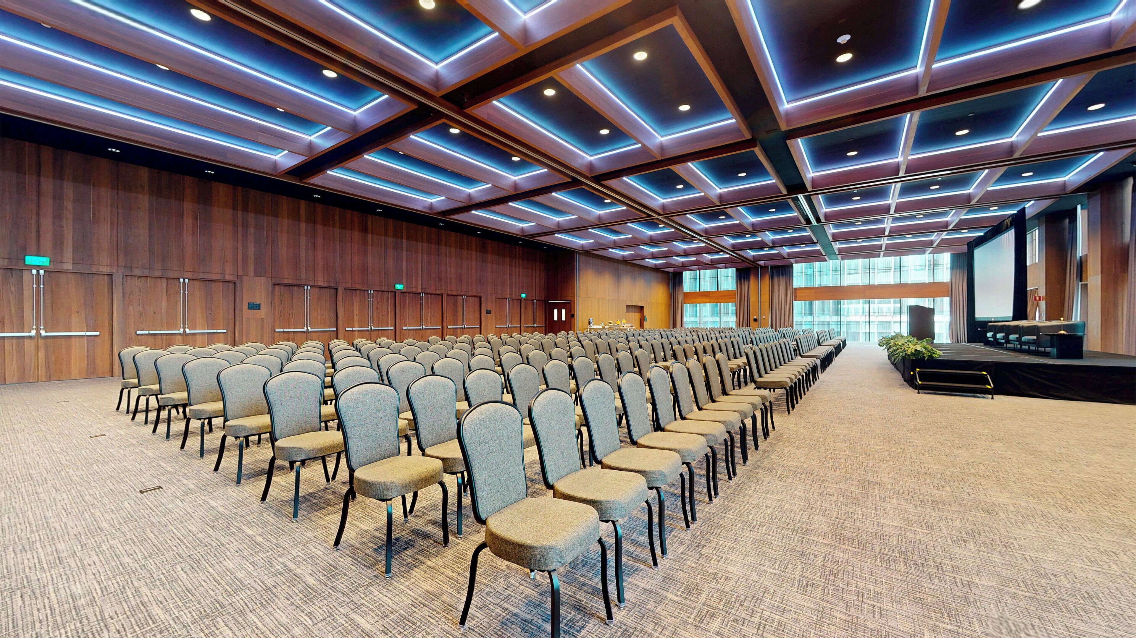 A large conference room with rows of chairs, a stage, and a wooden wall with doors.