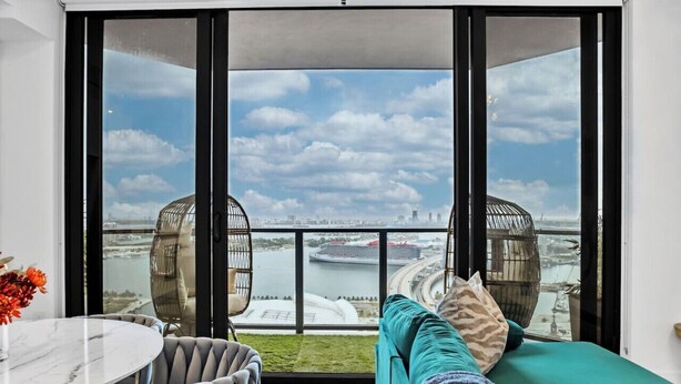 Un salon moderne avec un canapé, une table ronde et des chaises, offrant une vue sur un paysage urbain et une étendue d’eau grâce à de grandes fenêtres.