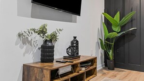 Un mueble de entretenimiento de madera con un televisor de pantalla plana montado encima. Hay dos plantas en macetas sobre el mueble, una de hojas anchas y otra con follaje más pequeño. También hay un control remoto y un libro titulado "Sex in the City" en el estante.
