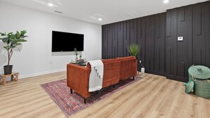 Een moderne woonkamer met een bank, een flatscreen televisie, een donkere houten wandbekleding, een klein tafeltje met een plant en een kamerkleed met een patroon.