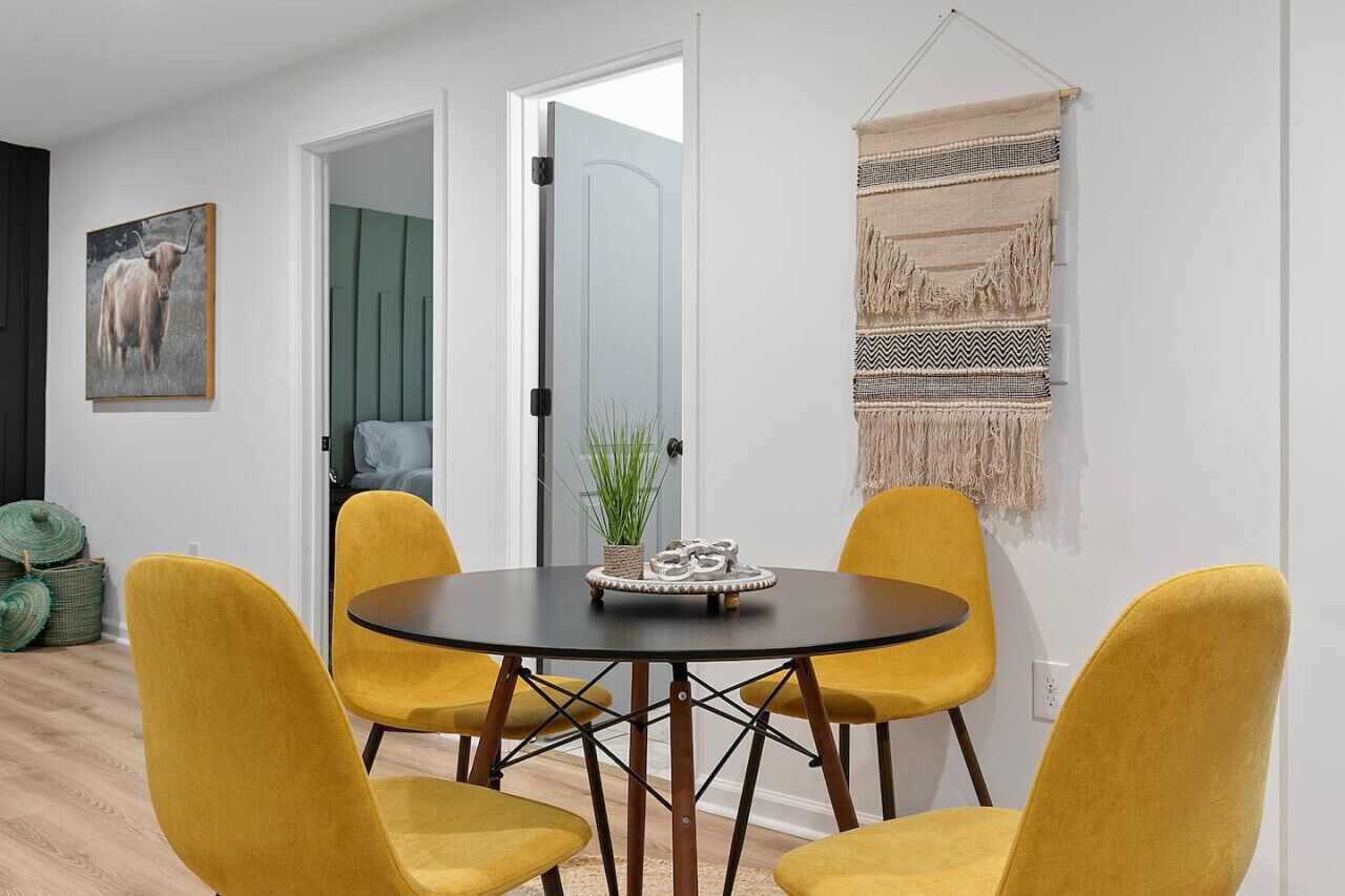 A dining area with a round table and four yellow chairs, a framed picture on the wall, and a woven tapestry.