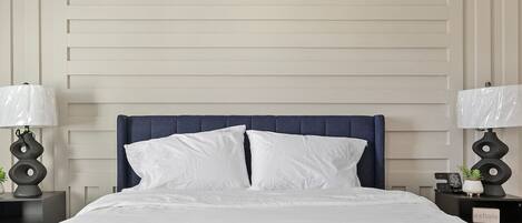 A neatly made bed with white linens, a dark blue headboard, and two bedside tables with lamps and small decorative items.