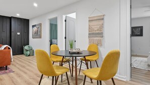 Een eetgedeelte met een ronde tafel en vier gele stoelen, een grijze bank en een donkere houten wand.