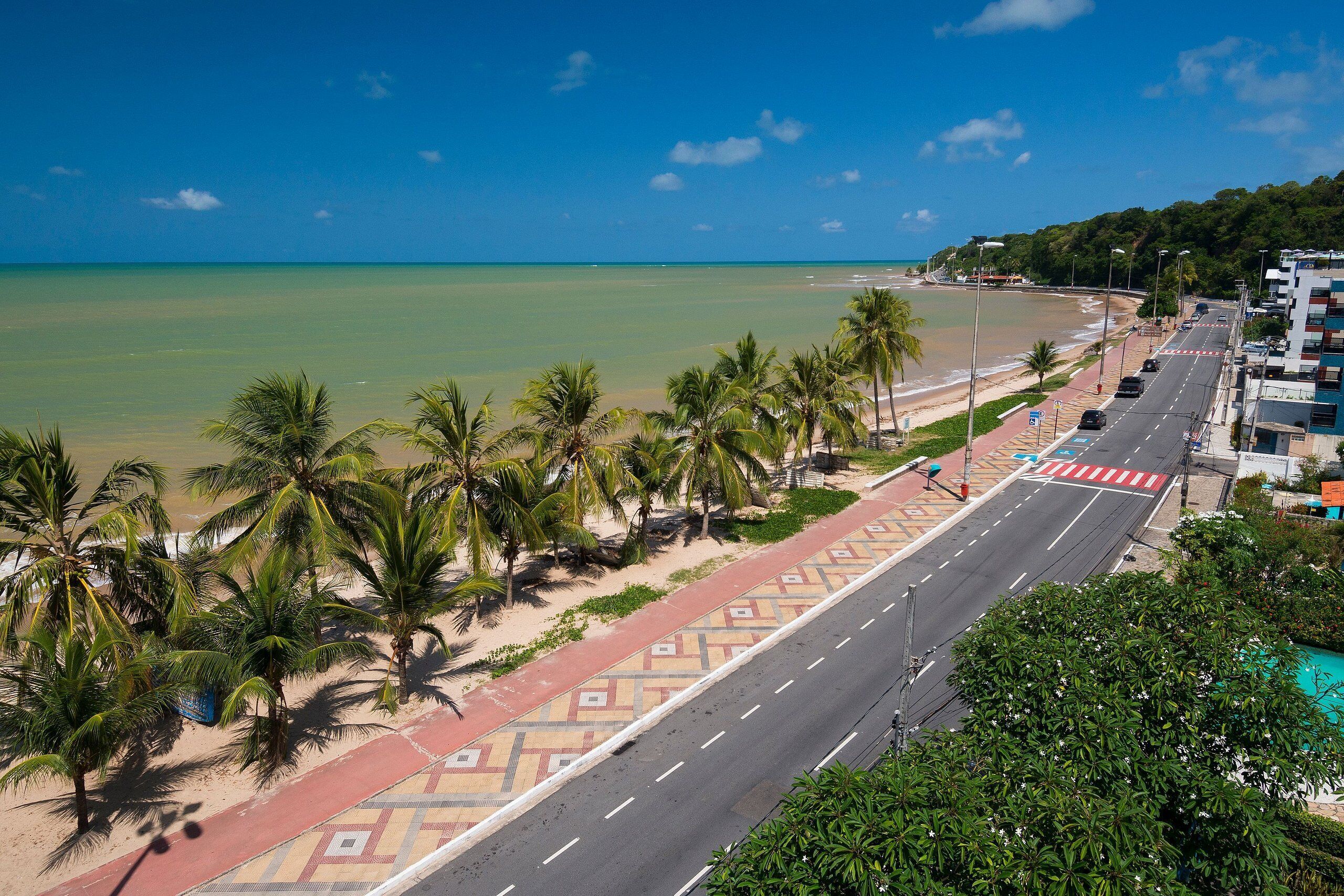 A coastal road with palm trees, a beach, and buildings.