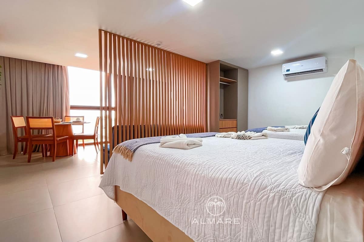 A hotel room with a bed, a desk, and a chair. There is a window with curtains, an air conditioning unit, and a wardrobe.