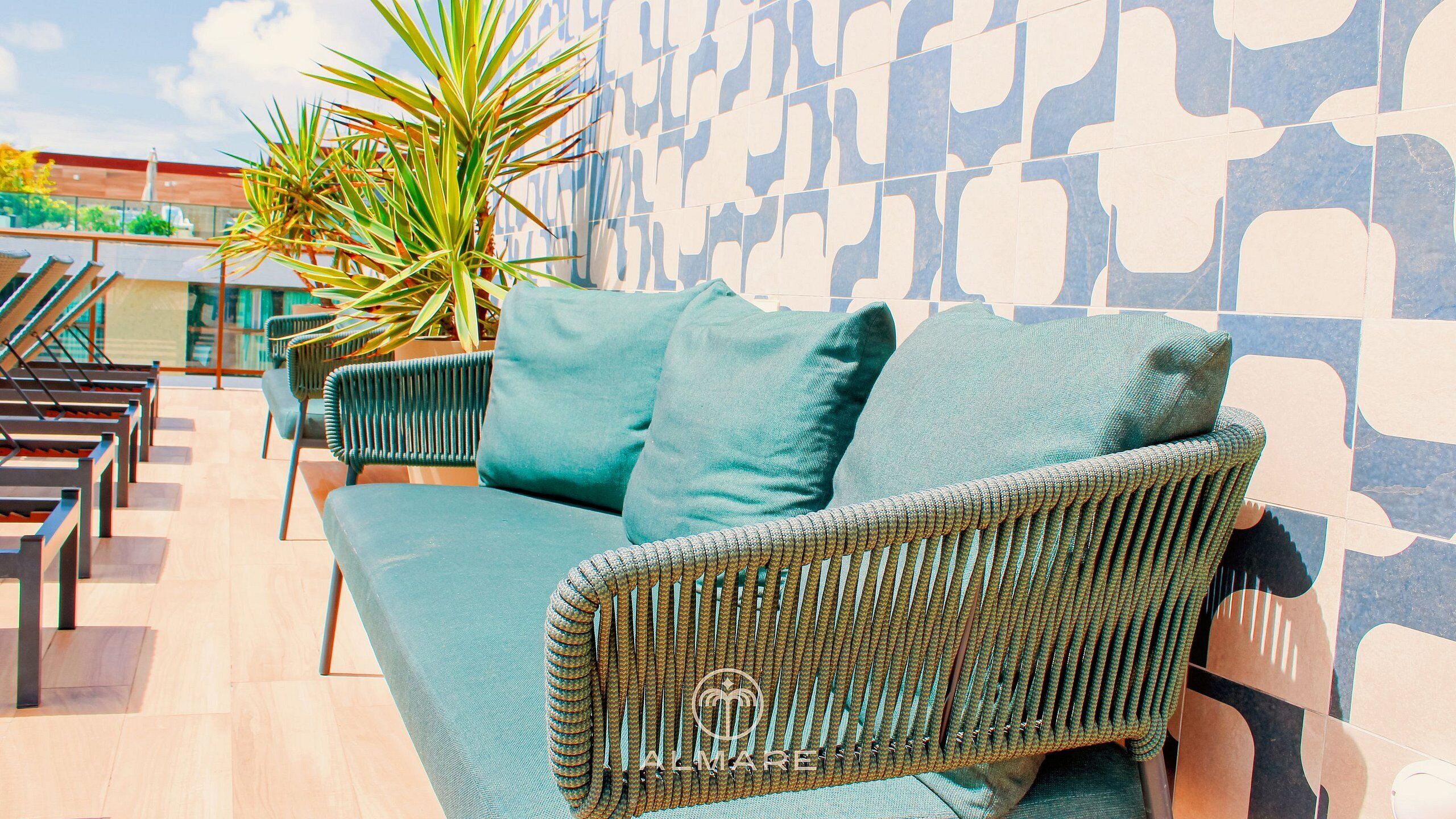 A wicker sofa with cushions, a potted plant, and a patterned wall in a sunny outdoor seating area.