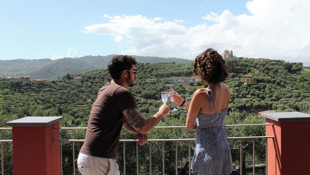 Un couple se tient sur un balcon, d’où l’on peut admirer un paysage pittoresque composé de collines et de bâtiments.