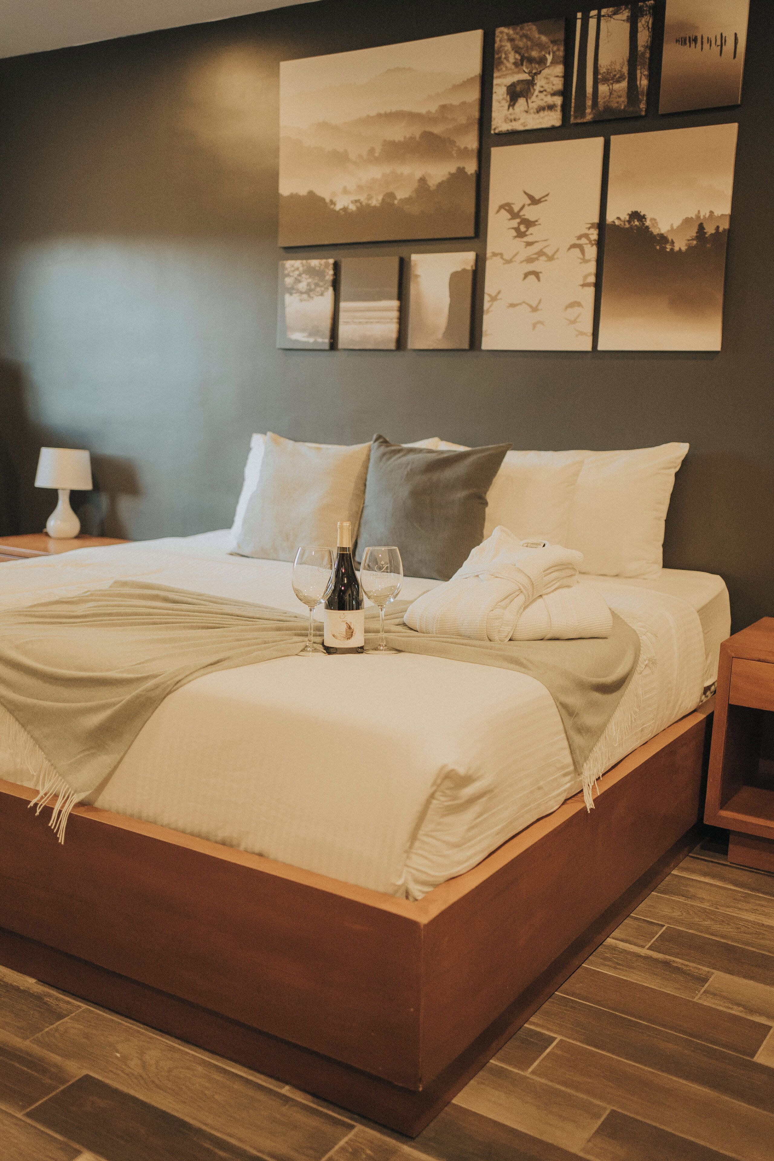 Una cama bien hecha con una botella y vasos, un cabecero de madera y una pared con cuadros.