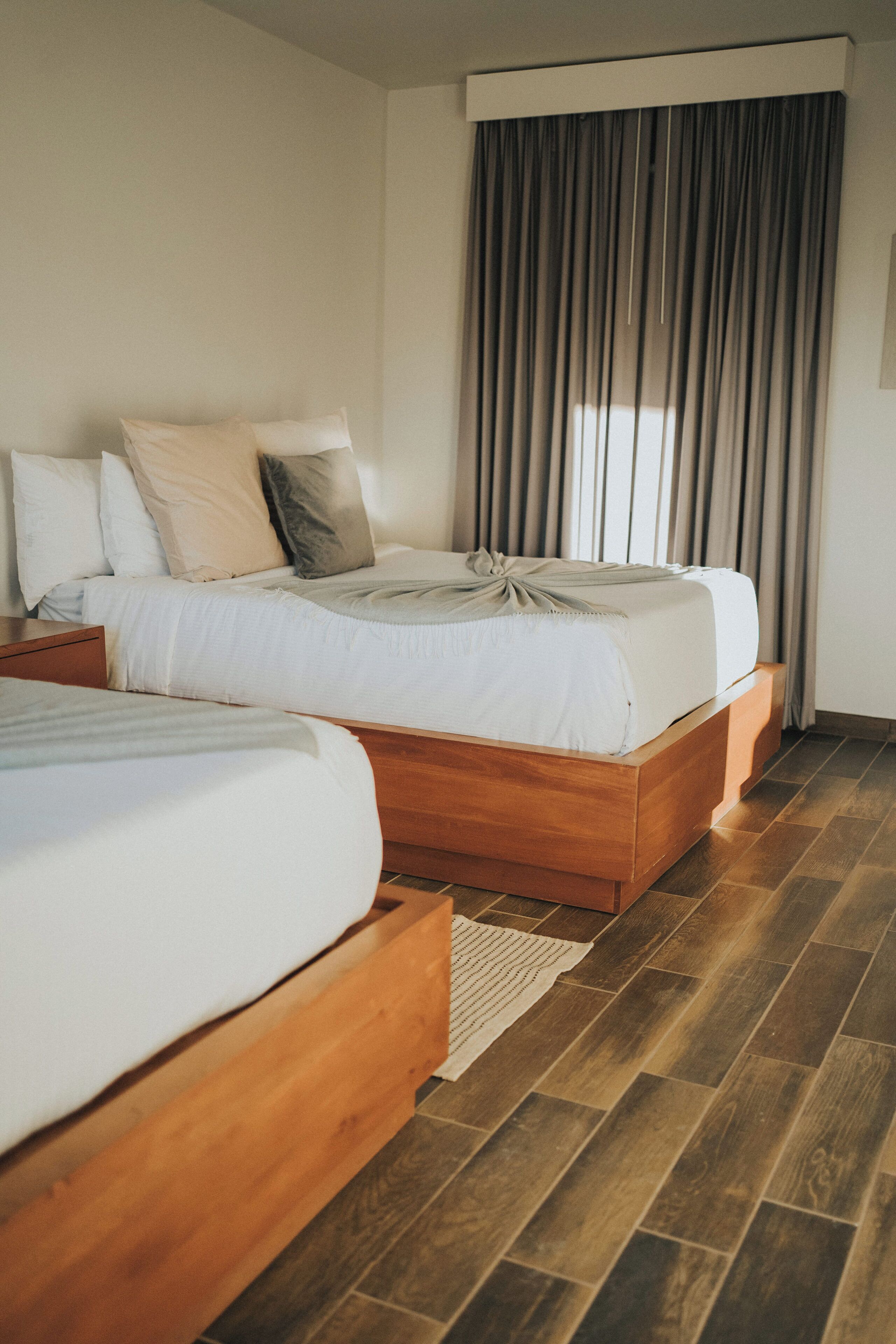 Quarto de hotel com duas camas de madeira, uma janela grande com cortinas e piso de madeira.
