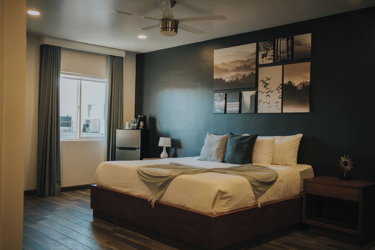 Une chambre moderne avec un grand lit, un ventilateur de plafond, une table de chevet et une œuvre d’art accrochée au mur.