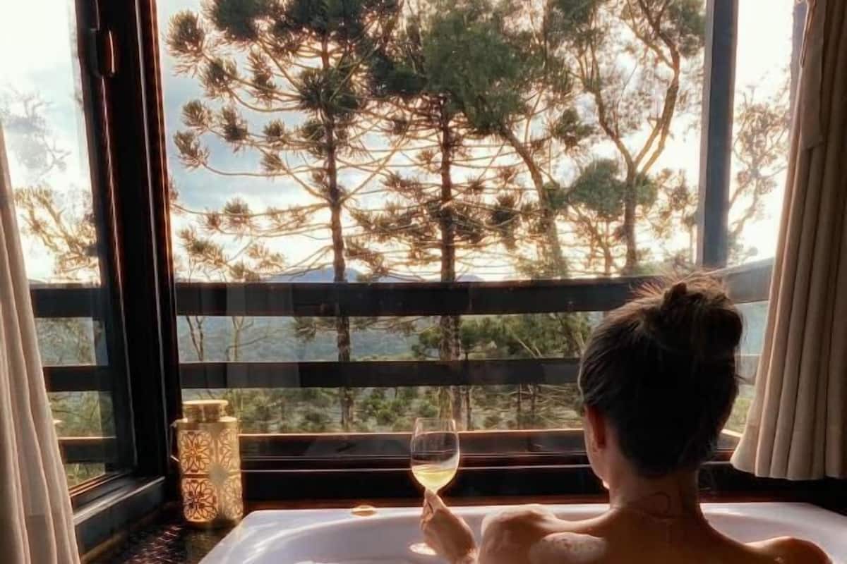 A person relaxing in a hot tub with a glass of wine, overlooking a view of trees through large windows.