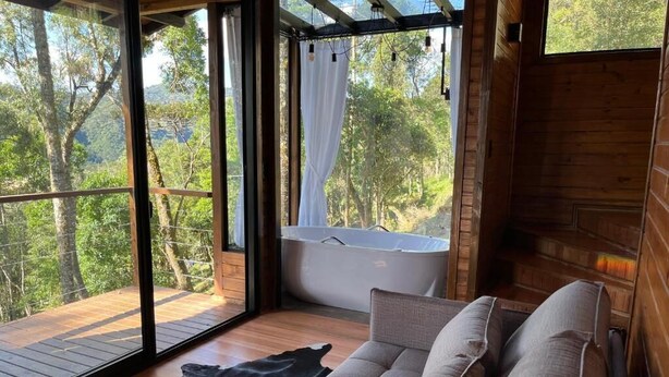 Una cabaña de madera con un gran ventanal de vidrio que da a una terraza, un sofá y una bañera.