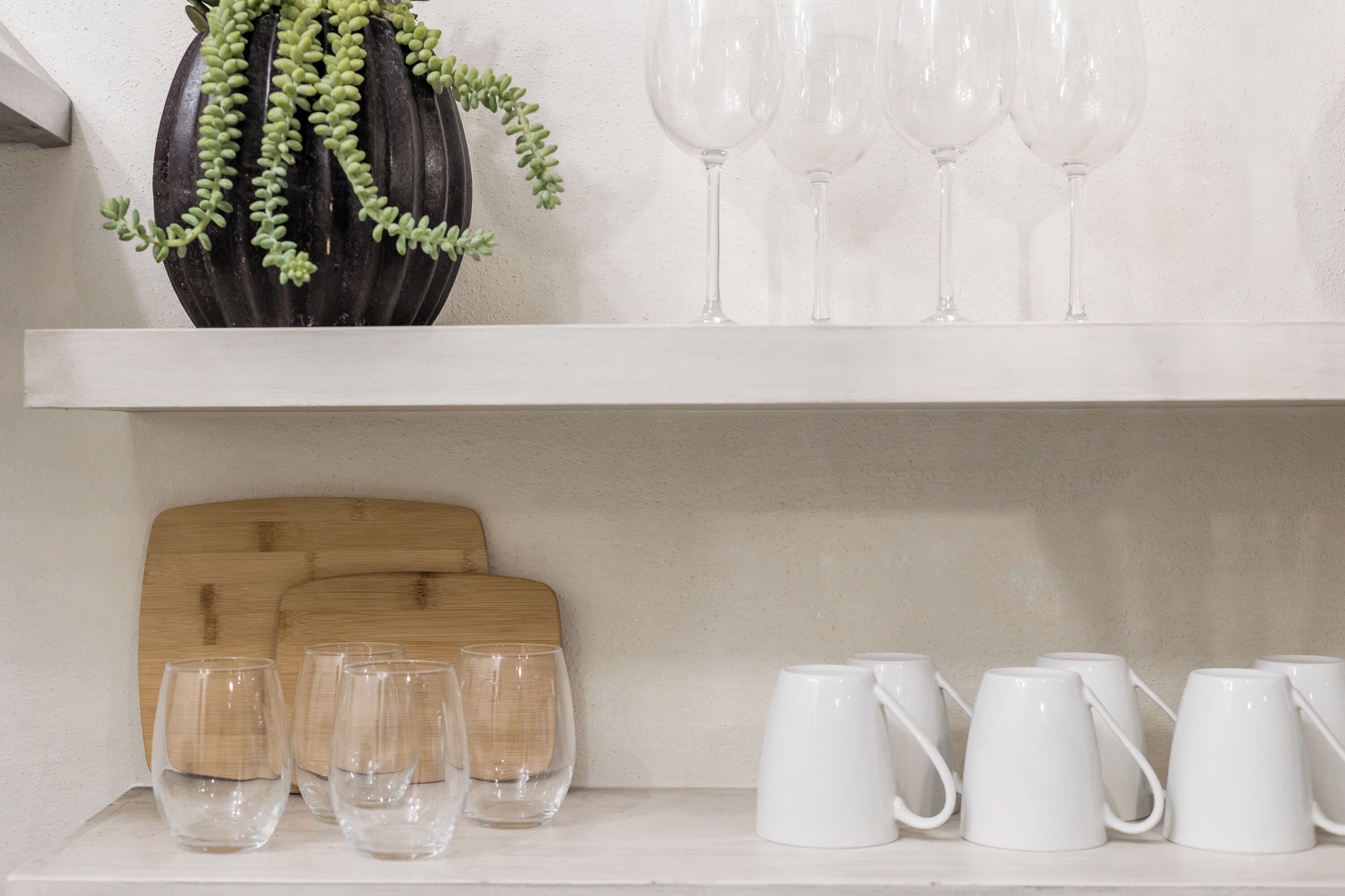 A shelf with a black vase with green plants, several wine glasses, and white mugs.