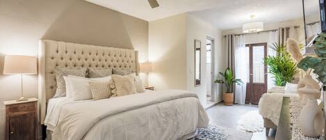 A bedroom with a tufted headboard, a large bed, a ceiling fan, and a view of the outdoors.
