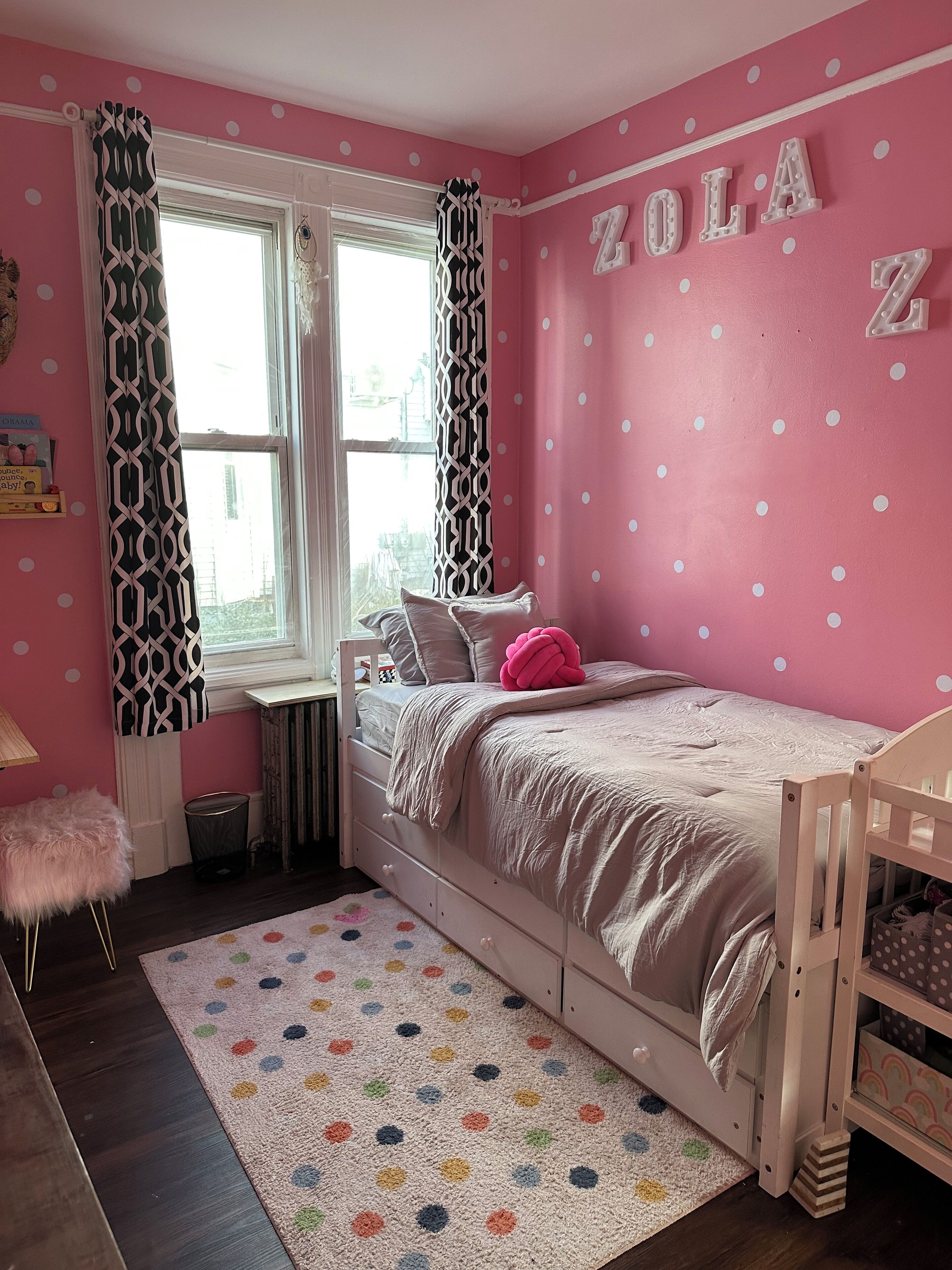 Une chambre à coucher décorée dans des tons roses, avec un lit, une fenêtre ornée de rideaux à motifs, un tapis à pois et un berceau.